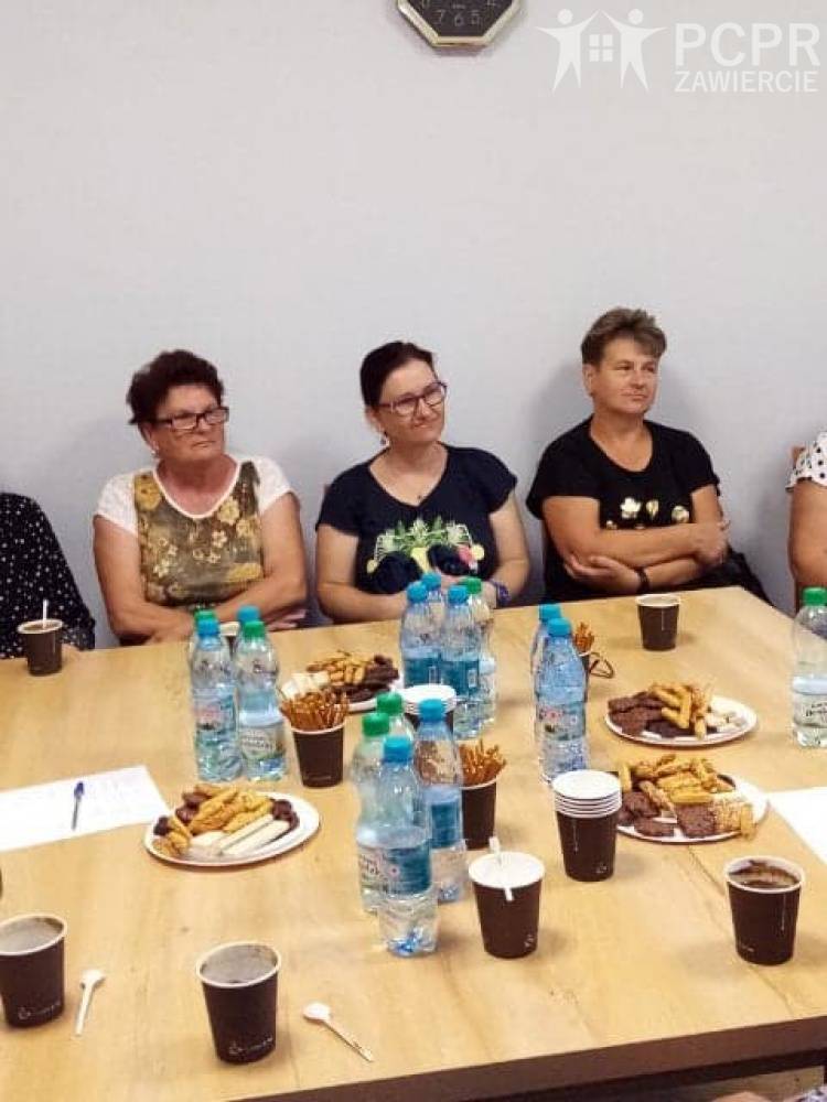 Zdjęcie: Grupa kobiet siedzi przy stole, na którym znajdują się ciastka, woda mineralna i kubeczki